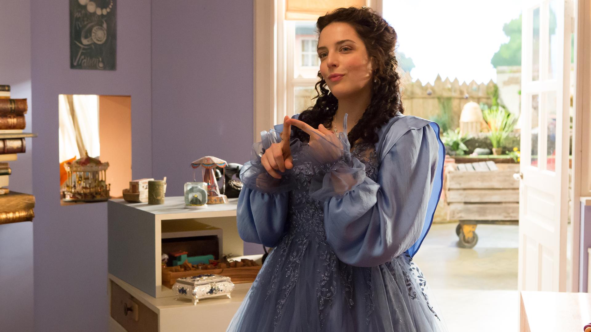 Siebenstein als Prinzessin, mit langem, schwarzen Haar und einem blauen Prinzessinnenkleid, steht im Laden und lächelt verschmitzt. Im Hintergrund sieht man die geöffnete Tür zum Hof hin und Regale mit allerlei Krims-Krams.