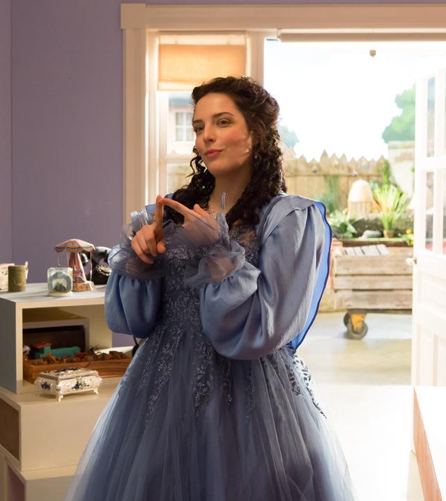 Siebenstein (Anna Werner Friedmann) als Prinzessin, mit langem, schwarzen Haar und einem blauen Prinzessinnenkleid, steht im Laden und lächelt verschmitzt. Im Hintergrund sieht man die geöffnete Tür zum Hof hin und Regale mit allerlei Krims-Krams. 