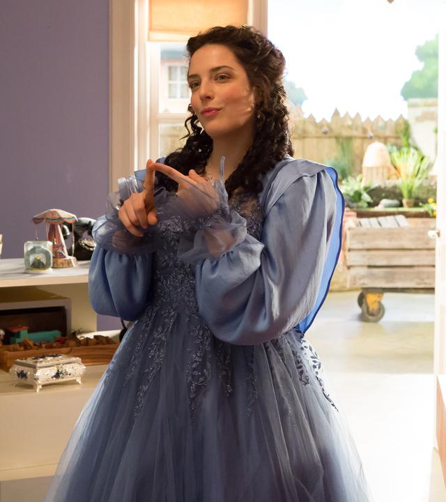 Siebenstein als Prinzessin, mit langem, schwarzen Haar und einem blauen Prinzessinnenkleid, steht im Laden und lächelt verschmitzt. Im Hintergrund sieht man die geöffnete Tür zum Hof hin und Regale mit allerlei Krims-Krams.