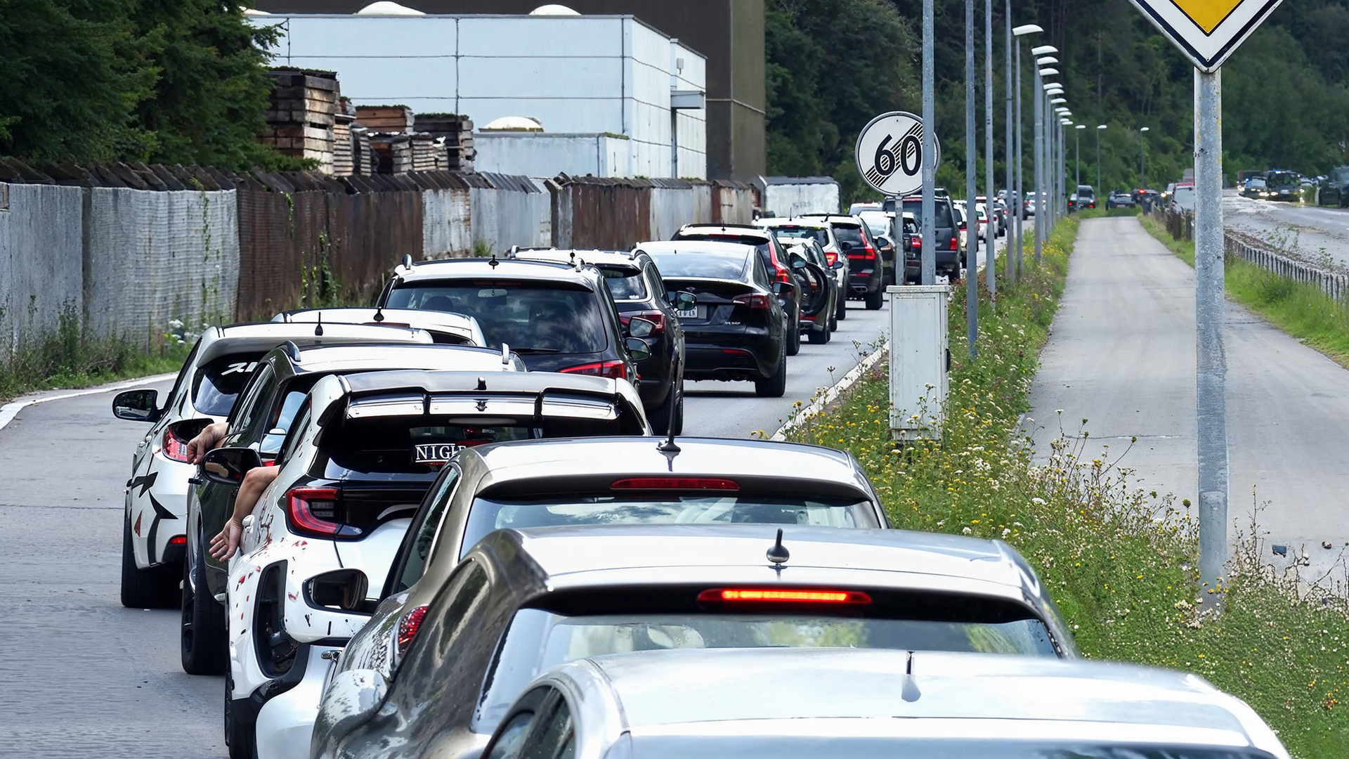 Langer Rückstau von Autos auf einer Straße neben einem Industriegelände. Einige Fahrer strecken ihre Arme aus den Autofenstern, während die Fahrzeuge dicht an dicht stehen. Rechts neben der Straße verläuft ein leerer Gehweg, gesäumt von Straßenlaternen und Wildblumen.