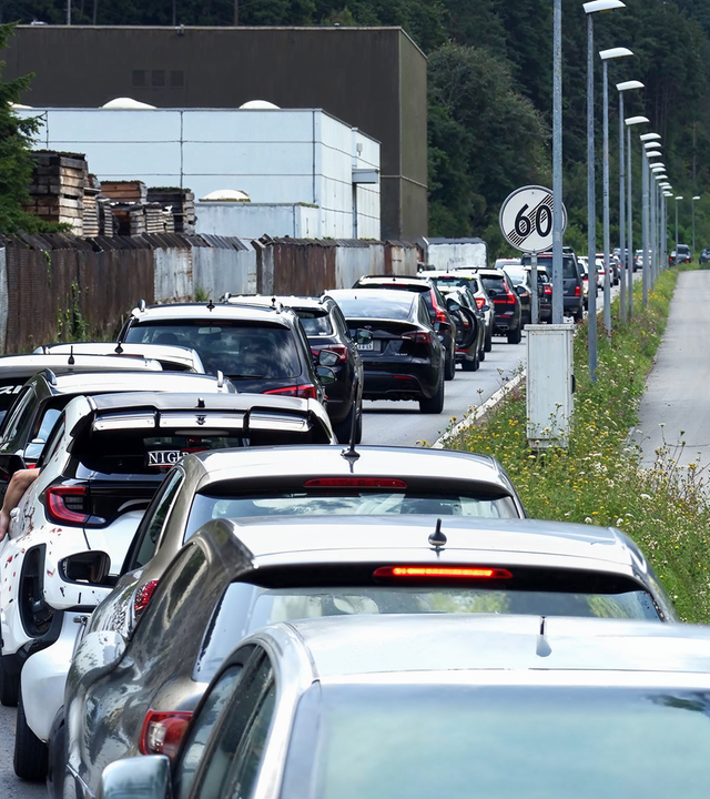 Langer Rückstau von Autos auf einer Straße neben einem Industriegelände. Einige Fahrer strecken ihre Arme aus den Autofenstern, während die Fahrzeuge dicht an dicht stehen. Rechts neben der Straße verläuft ein leerer Gehweg, gesäumt von Straßenlaternen und Wildblumen.