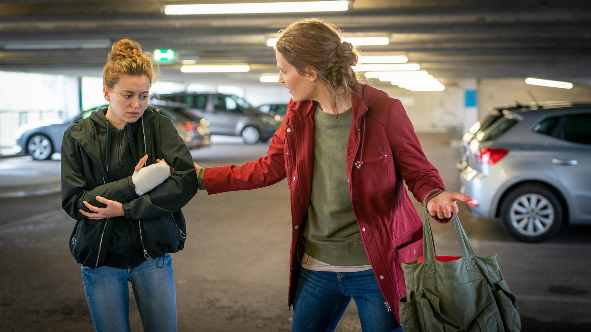 "Notruf Hafenkante - Überlebenskampf": Pola (Alexandra Pfeifer) und ihre Mutter Lena (Nora Moltzen) stehen in einem Parkhaus nebeneinander. Lena zieht ihre Tochter am Arm. Sie hat sich zu ihr gedreht. Pola blickt nachdenklich ins Leere. Sie hält sich ihren rechten eingegipsten Arm.
