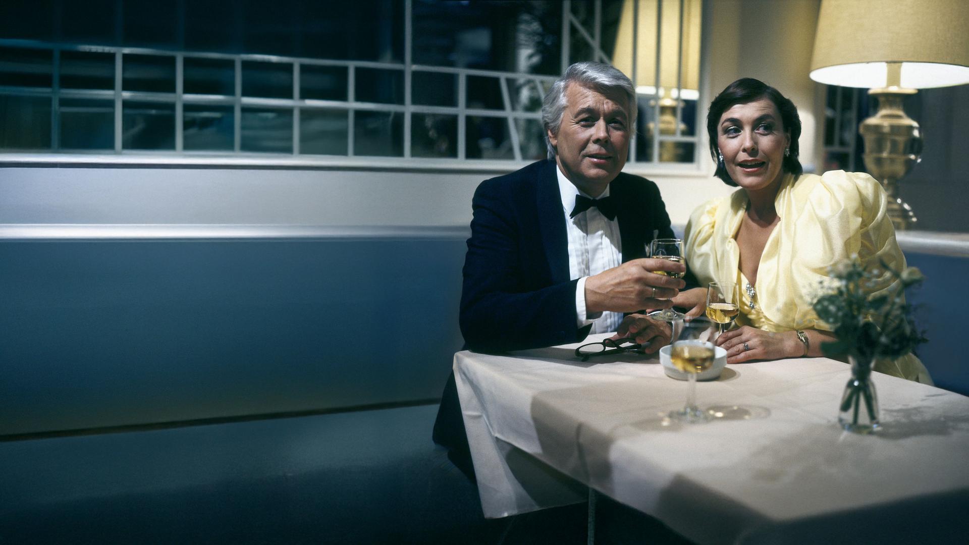 Werner und Angi sitzen in festlicher Kleidung an einem Tisch und halten jeweils beide ein Weinglas in der Hand. Beide blicken in dieselbe Richtung und schauen freudig erstaunt.