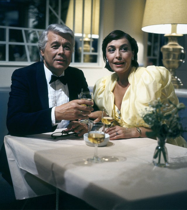 Werner und Angi sitzen in festlicher Kleidung an einem Tisch und halten jeweils beide ein Weinglas in der Hand. Beide blicken in dieselbe Richtung und schauen freudig erstaunt.