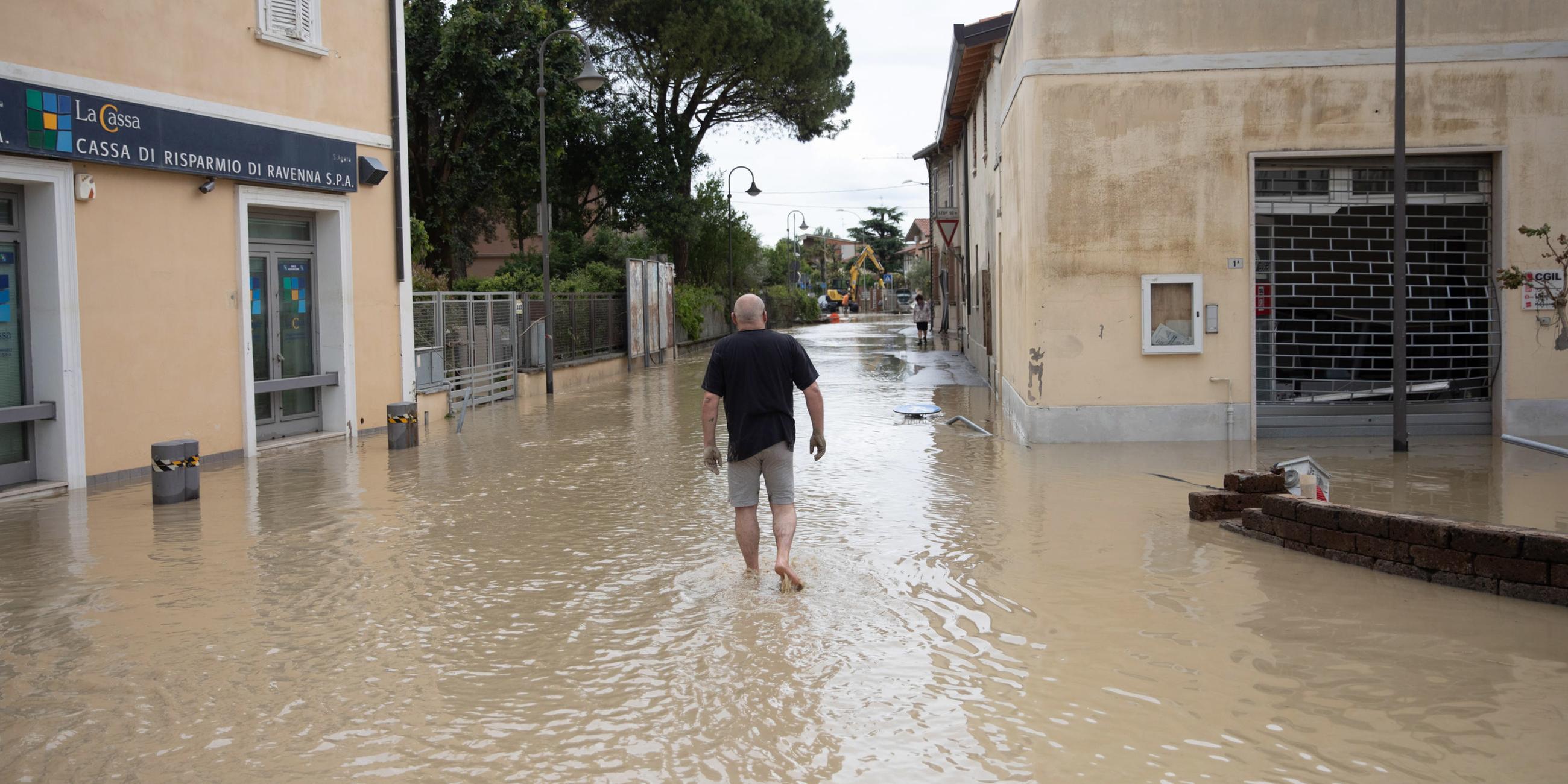 Zu sehen ist ein Mann, der durch dreckiges Wasser zwischen Häusern in der Region Emilia-Romagna watet.