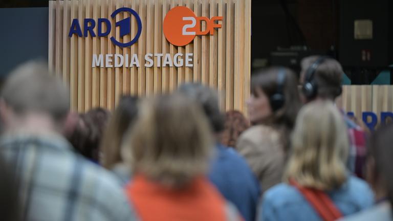 Die „ARD ZDF Media Stage“ ist als Schriftzug auf einer vertikalen Holzpaneel-Wand zu sehen. Davor befindet sich eine Menge von Besucherinnen und Besuchern, deren Köpfe und Schultern unscharf im Vordergrund erkennbar sind.
