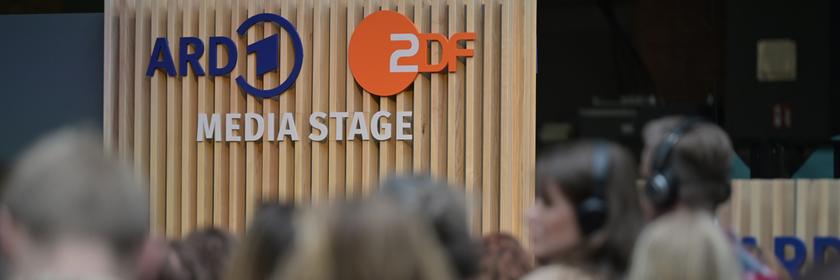 Die „ARD ZDF Media Stage“ ist als Schriftzug auf einer vertikalen Holzpaneel-Wand zu sehen. Davor befindet sich eine Menge von Besucherinnen und Besuchern, deren Köpfe und Schultern unscharf im Vordergrund erkennbar sind.