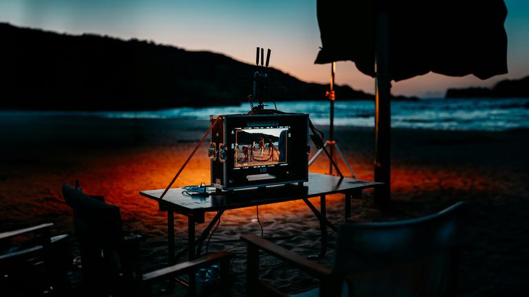 Ein Bildschirm in einem Produktionskoffer auf einem Tisch vor einem Strand, der in schönem Sonnenuntergangslicht liegt. Typical für Filmproduktionen