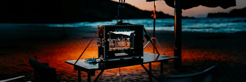 Ein Bildschirm in einem Produktionskoffer auf einem Tisch vor einem Strand, der in schönem Sonnenuntergangslicht liegt. Typical für Filmproduktionen