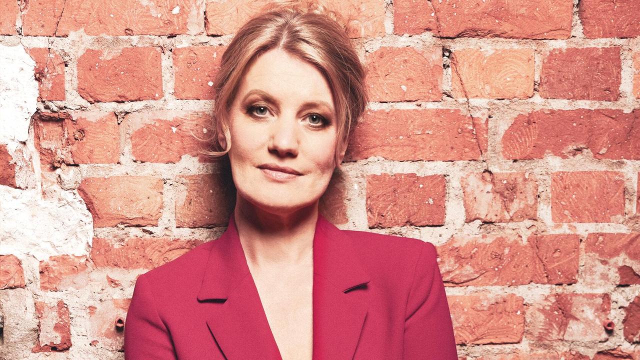 Die Sängerin Ulita Knaus in rotem Anzug, auf dem Boden sitzend, direkt in die Kamera schauend, an eine Ziegelwand gelehnt.