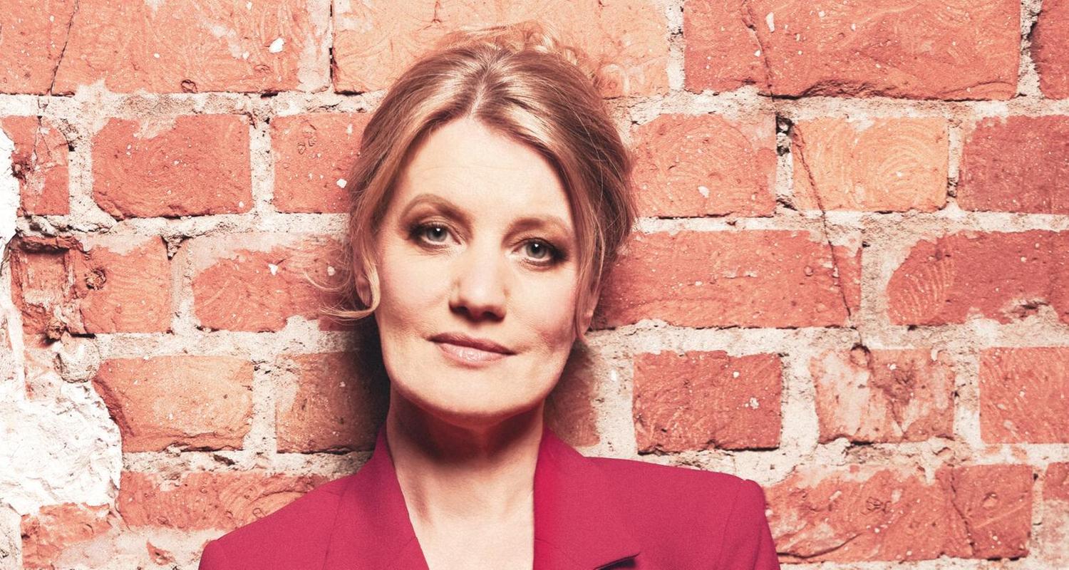 Die Sängerin Ulita Knaus in rotem Anzug, auf dem Boden sitzend, direkt in die Kamera schauend, an eine Ziegelwand gelehnt.
