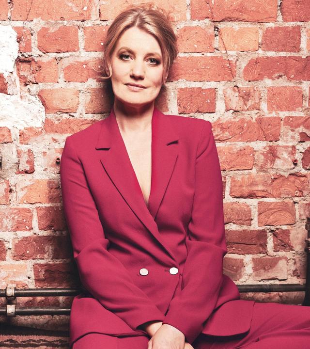 Die Sängerin Ulita Knaus in rotem Anzug, auf dem Boden sitzend, direkt in die Kamera schauend, an eine Ziegelwand gelehnt.