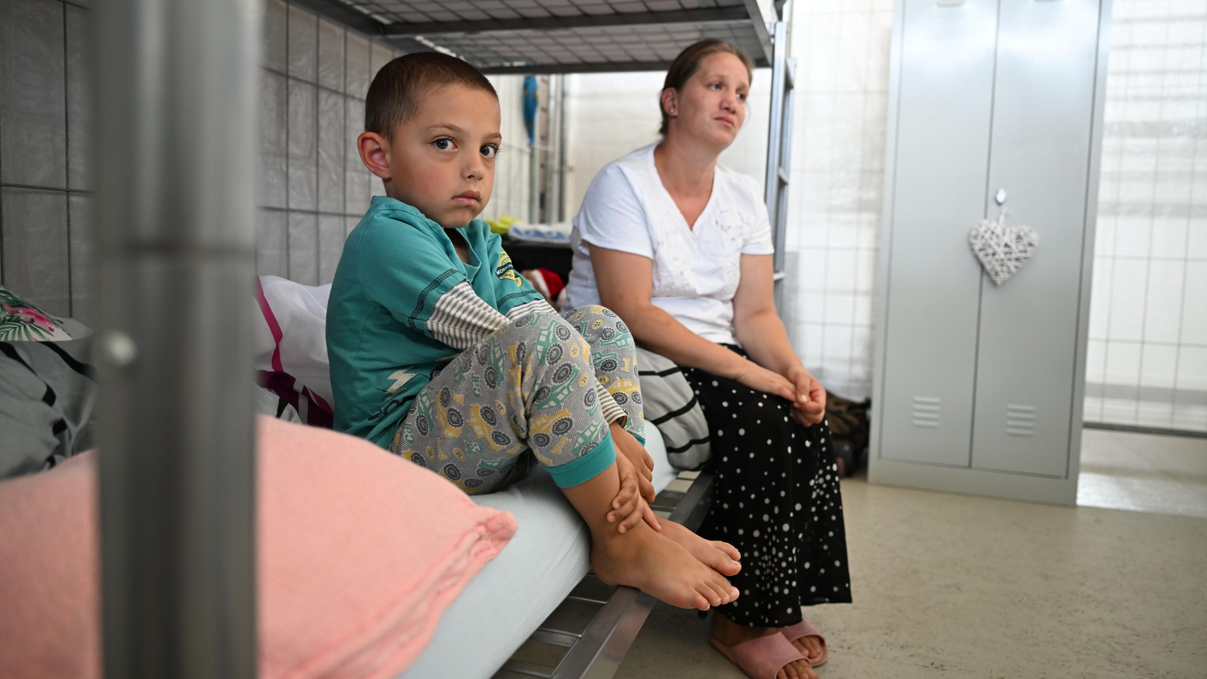 Eine Mutter und ihr Sohn sitzen auf einer Matratze in einem Stockbett im Zimmer einer Flüchtlingsunterkunft.
