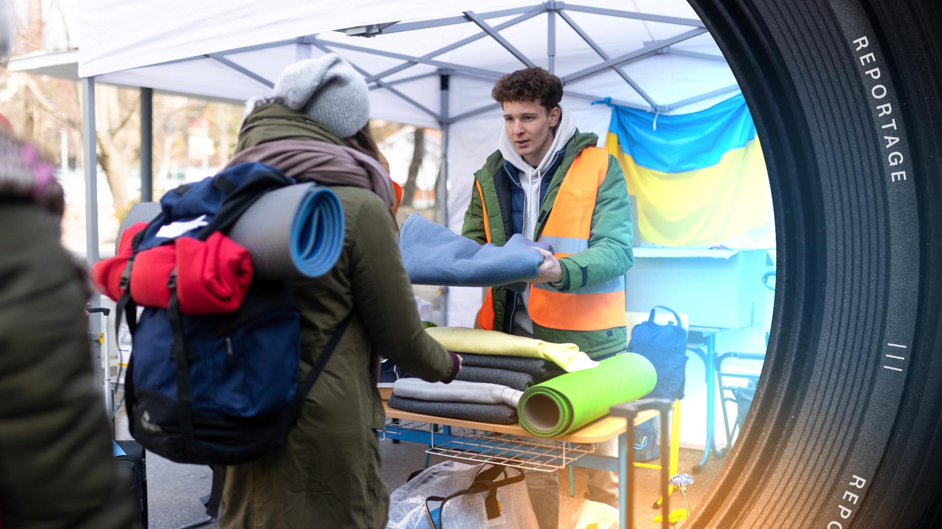 Ein junger Mann unter einem Zelt übergibt einer Frau mit großem Rucksack eine blaue Decke. Im Hintergrund eine große Ukraine-Flagge.