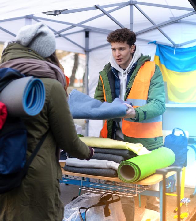 Ein junger Mann unter einem Zelt übergibt einer Frau mit großem Rucksack eine blaue Decke. Im Hintergrund eine große Ukraine-Flagge.