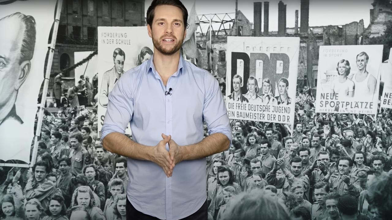 Mirko Drotschmann vor einem Foto mit einem Menschenzug mit DDR-Plakaten