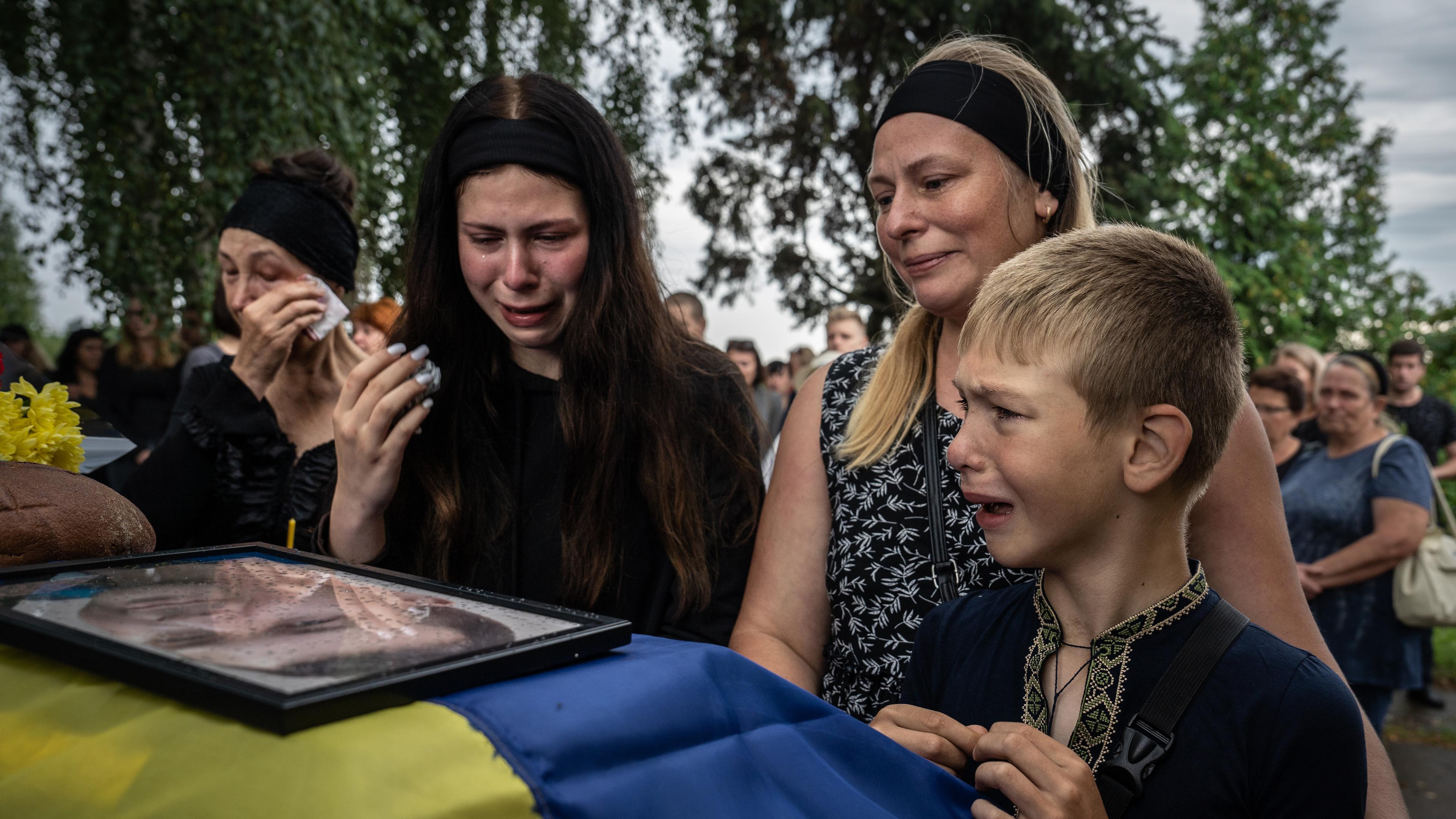 Zwei Frauen und ein kleiner Junge stehen um einen Sarg, auf dem die ukrainische Flagge und ein gerahmtes Bild liegen.