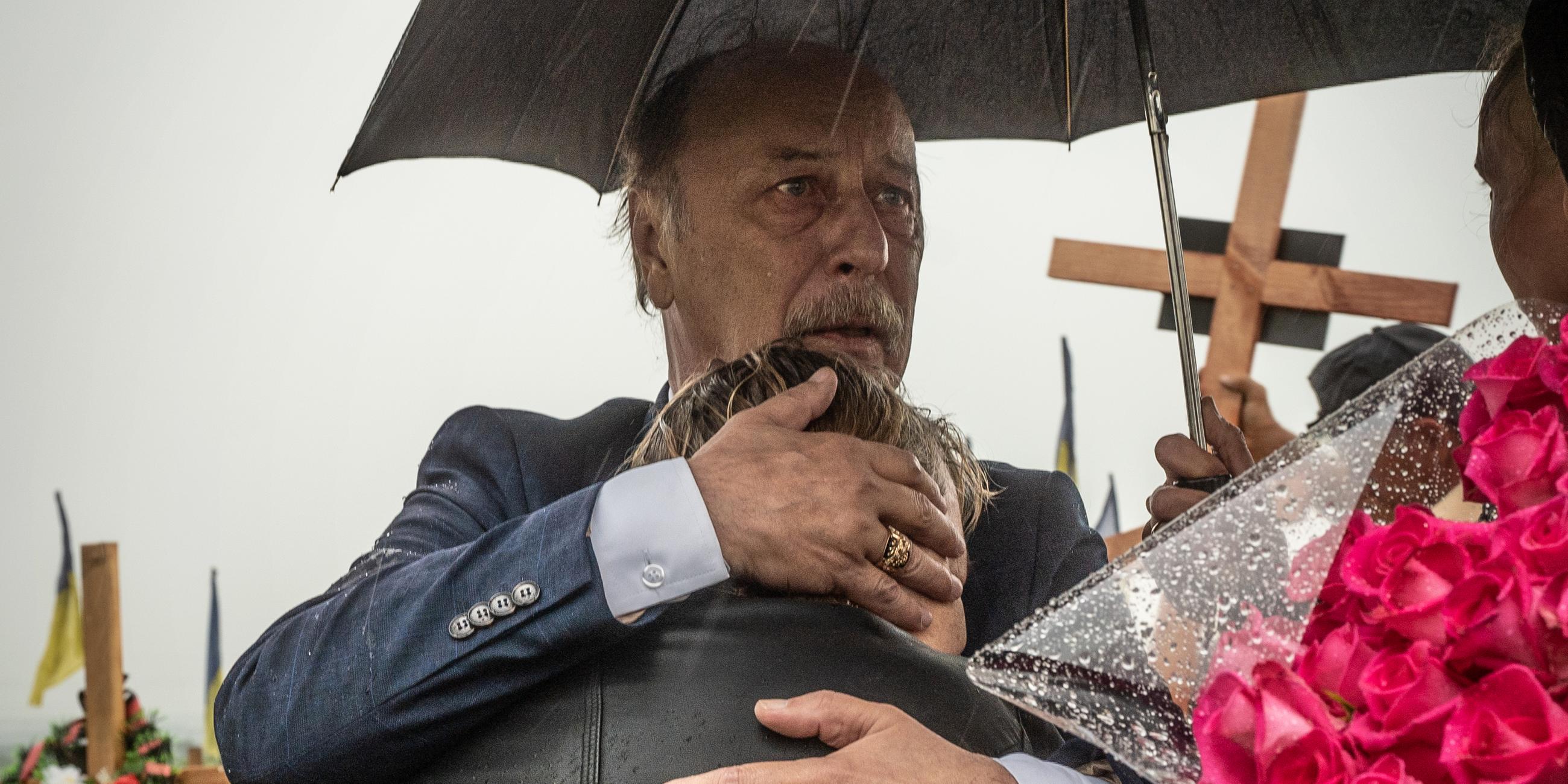 Ein Mann hält eine Frau in den Armen, über ihnen ein Regenschirm, im Hintergrund ist ein Friedhof zu sehen.
