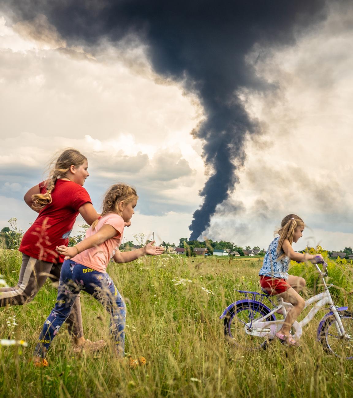 Drei Mädchen fahren Fahrrad in einem Feld, dahinter ist eine schwarze Rauchwolke zu sehen.