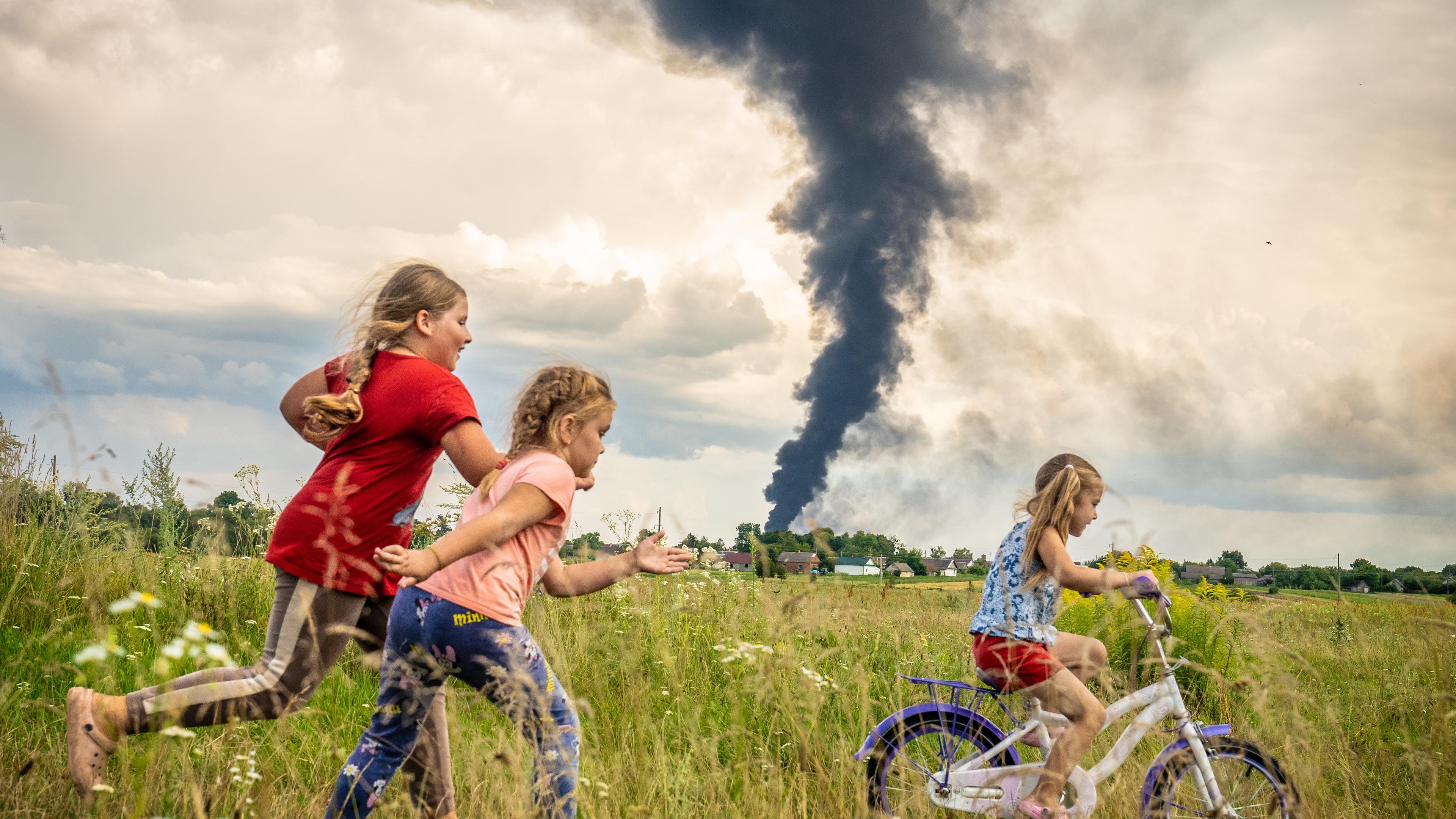 Drei Mädchen fahren Fahrrad in einem Feld, dahinter ist eine schwarze Rauchwolke zu sehen.
