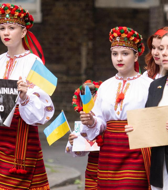 Drei junge Mädchen in ukrainischer Tracht und ukrainischen Fahnen in der Hand laufen über die Straße. Ein Mädchen trägt ein Schild mit der Aufschrift: Free Ukrainian Children Kidnapped by Russia. Mit dabei ist eine Frau, die einen braunen Umschlag in der Hand hält.