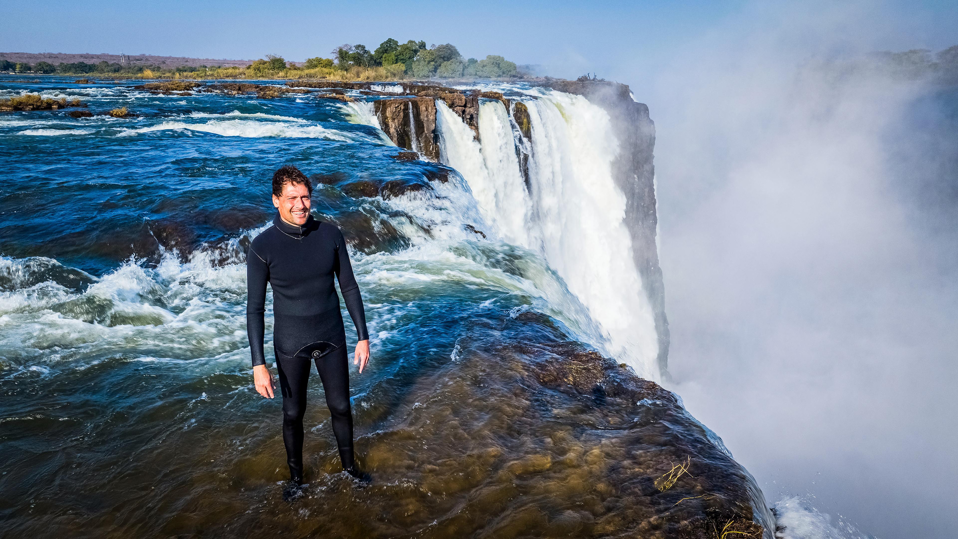 Moderator Uli Kunz steht im Taucheranzug auf der Abbruchkante der Viktoriafälle und lächelt in die Kamera.