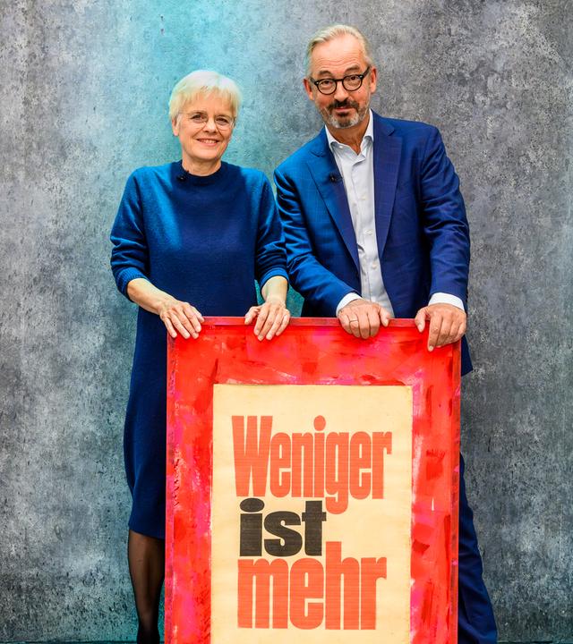 Jan Fleischhauer und Ulrike Herrmann stehen hinter einem Schild auf dem steht: Weniger ist mehr.