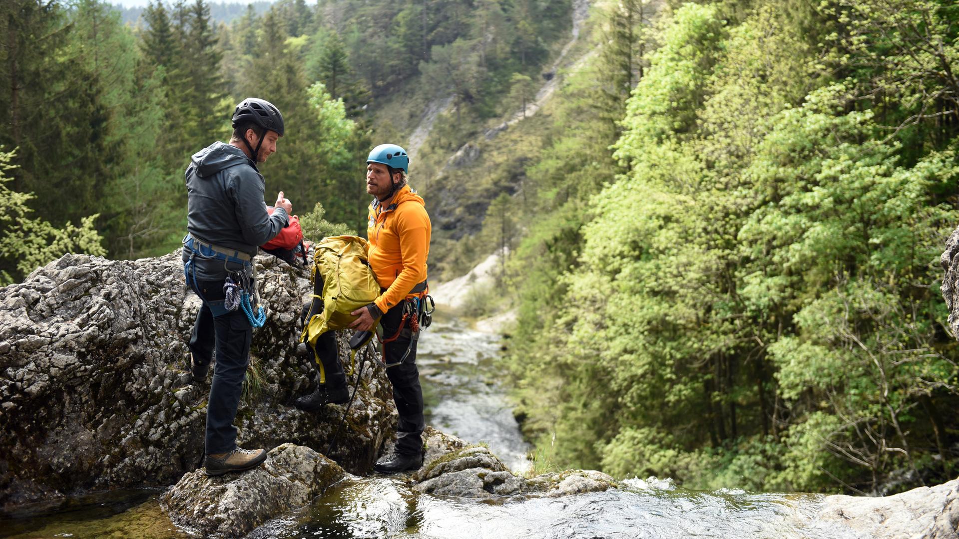 "Die Bergretter: Um jeden Preis": An einer Wasserfallklippe stehen sich Georg Haas (Leopold Hornung)und Bergretter Markus (Sebastian Ströbel) gegenüber. Sie tragen Helme und Kletterausrüstungen. Markus hält einen Rucksack in den Händen. Im Hintergrund ein bewaldeter Berghang.
