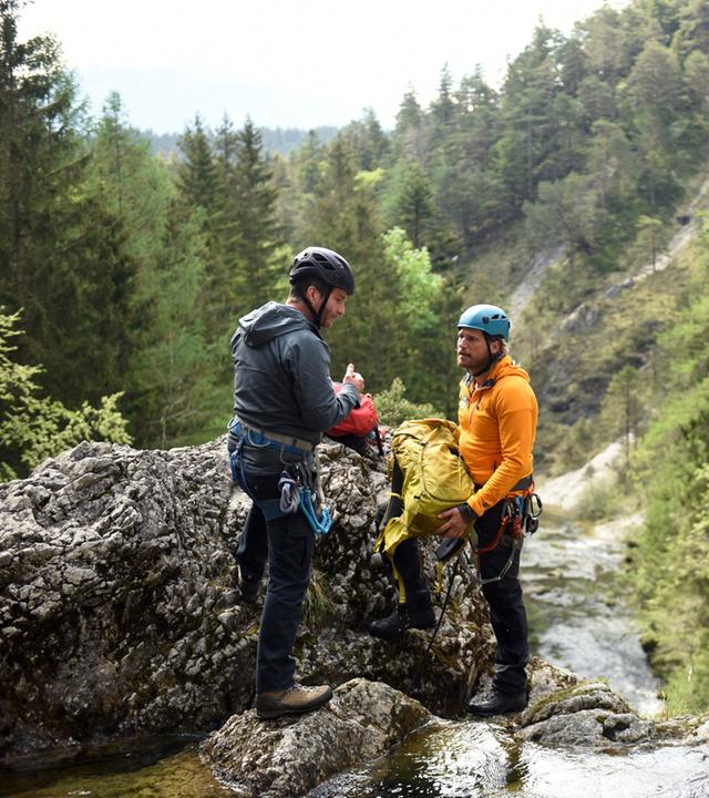 "Die Bergretter: Um jeden Preis": An einer Wasserfallklippe stehen sich Georg Haas (Leopold Hornung)und Bergretter Markus (Sebastian Ströbel) gegenüber. Sie tragen Helme und Kletterausrüstungen. Markus hält einen Rucksack in den Händen. Im Hintergrund ein bewaldeter Berghang.