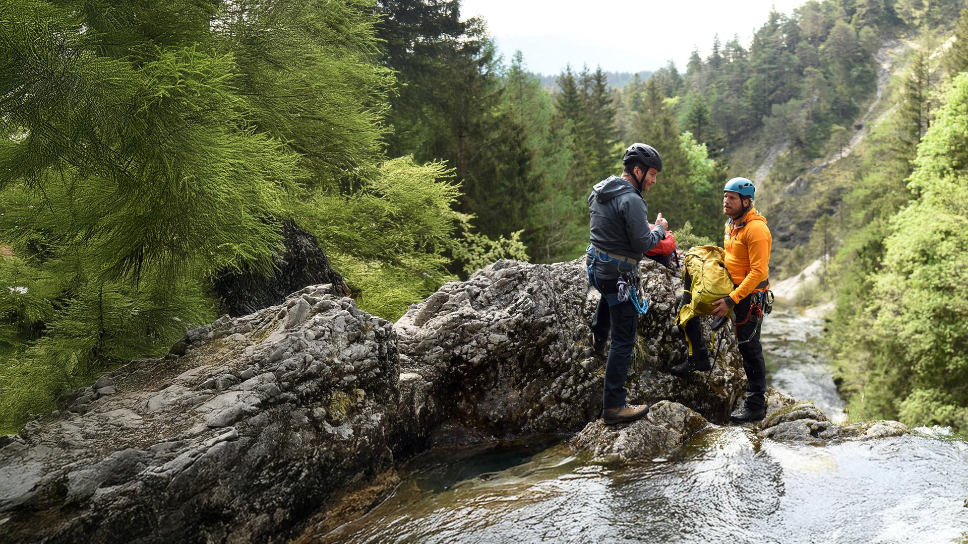 "Die Bergretter: Um jeden Preis": An einer Wasserfallklippe stehen sich Georg Haas (Leopold Hornung)und Bergretter Markus (Sebastian Ströbel) gegenüber. Sie tragen Helme und Kletterausrüstungen. Markus hält einen Rucksack in den Händen. Im Hintergrund ein bewaldeter Berghang.