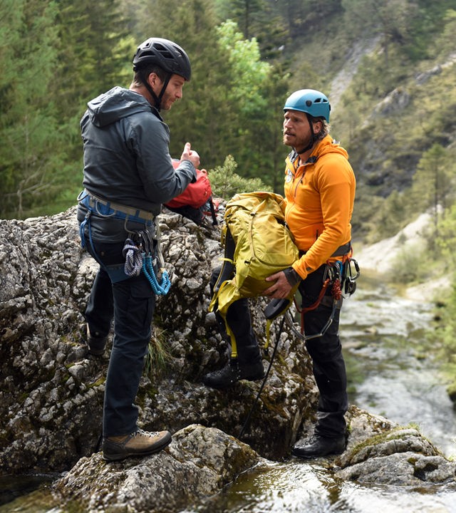 "Die Bergretter: Um jeden Preis": An einer Wasserfallklippe stehen sich Georg Haas (Leopold Hornung)und Bergretter Markus (Sebastian Ströbel) gegenüber. Sie tragen Helme und Kletterausrüstungen. Markus hält einen Rucksack in den Händen. Im Hintergrund ein bewaldeter Berghang.