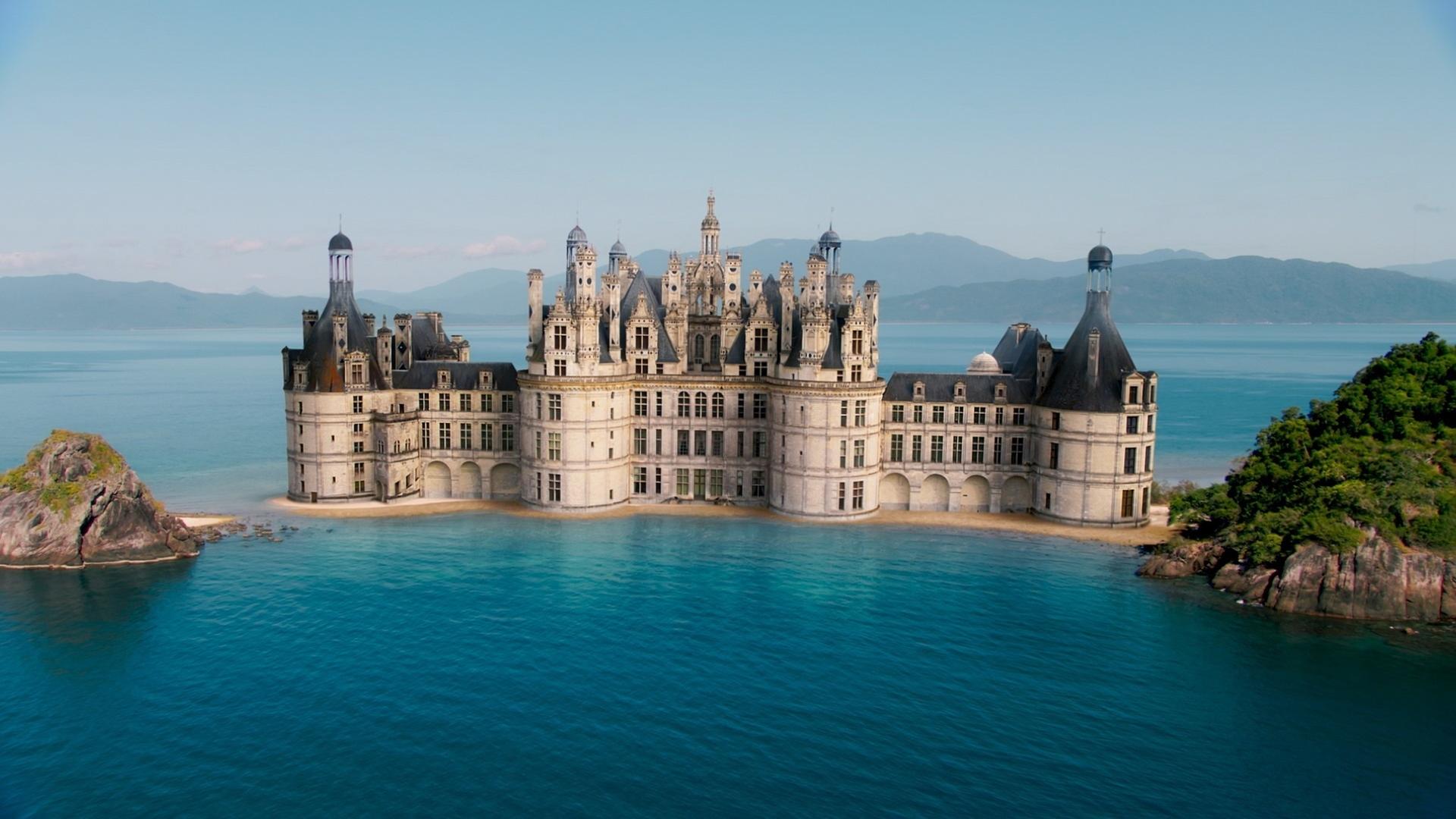 Abbildung eines Schlosses mit vielen großen und kleinen Türmen, umgeben von Wasser. Im Hintergrund befinden sich Berge, an den Seiten höhere bewachsene Steinhügel.
