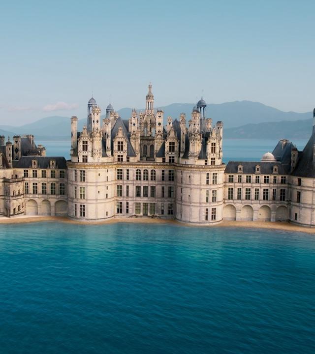 Abbildung eines Schlosses mit vielen großen und kleinen Türmen, umgeben von Wasser. Im Hintergrund befinden sich Berge, an den Seiten höhere bewachsene Steinhügel.