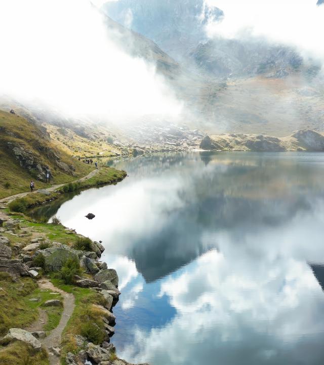 Eine malerische Berglandschaft im Piemont mit einem klaren See, dessen Wasser die umliegenden Berge und den Himmel mit Wolken spiegelt. Ein schmaler Wanderweg schlängelt sich am Ufer entlang, auf dem Wanderer zu sehen sind.