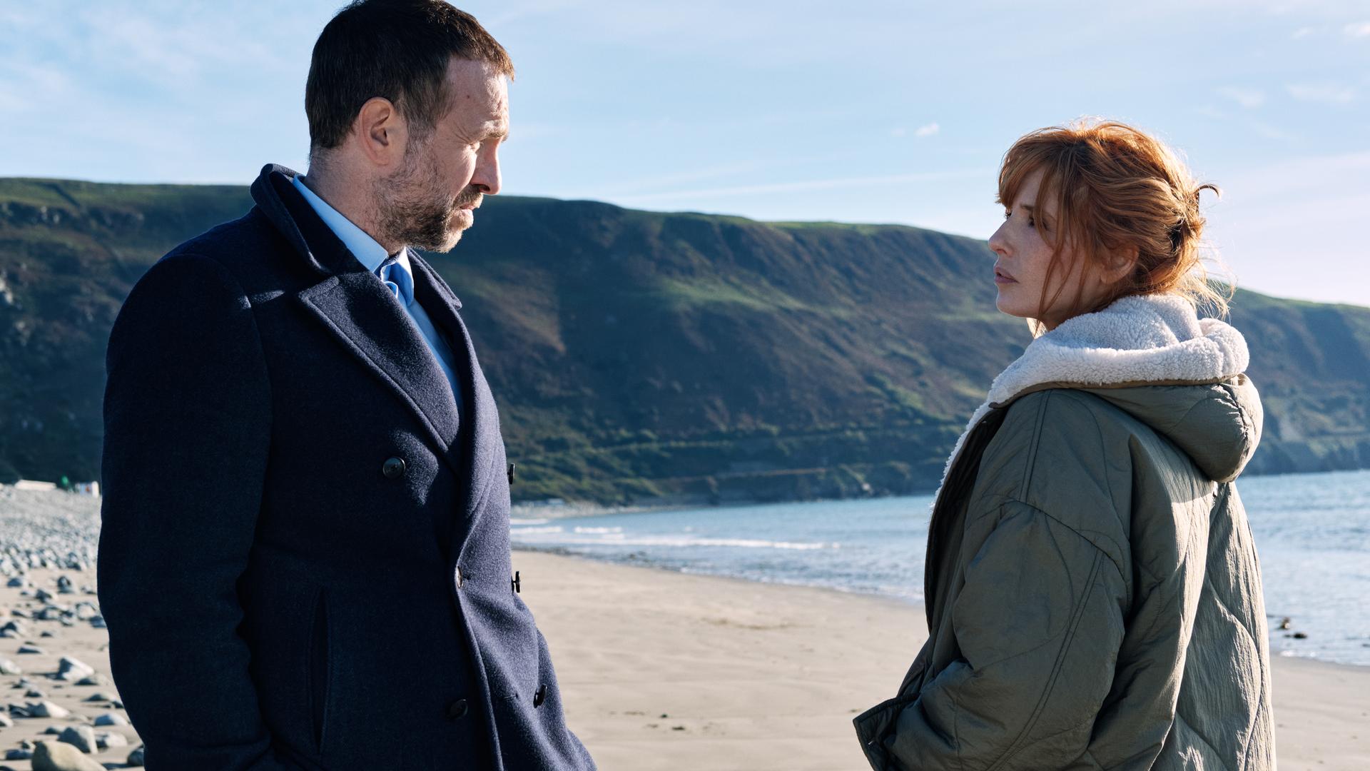 "Under Salt Marsh - Tod in den Salzwiesen - Ein grauenhafter Fund": Jackie Ellis (Kelly Reilly) und Detective Eric Bull (Rafe Spall) stehen sich an einem Strand gegenüber. Hinter ihnen erstrecken sich das Meer, der Sandstrand und grüne Steilklippen unter blauem Himmel.