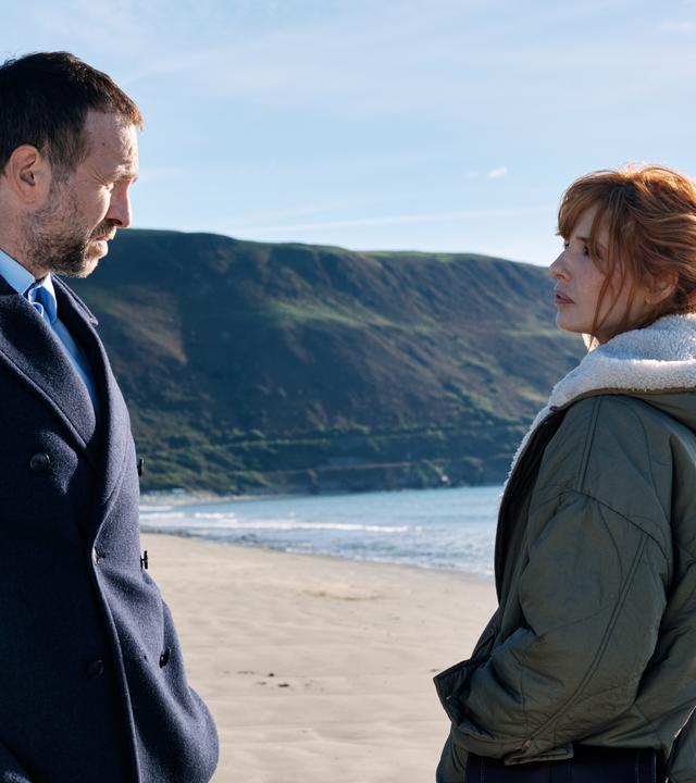 "Under Salt Marsh - Tod in den Salzwiesen - Ein grauenhafter Fund": Jackie Ellis (Kelly Reilly) und Detective Eric Bull (Rafe Spall) stehen sich an einem Strand gegenüber. Hinter ihnen erstrecken sich das Meer, der Sandstrand und grüne Steilklippen unter blauem Himmel.