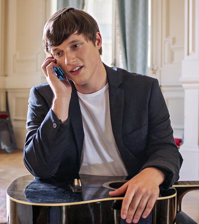 Henri sitzt auf einem Stuhl, seine Gitarre liegt auf seinem Schoß. Mit der rechten Hand hält er sein Handy ans Ohr.