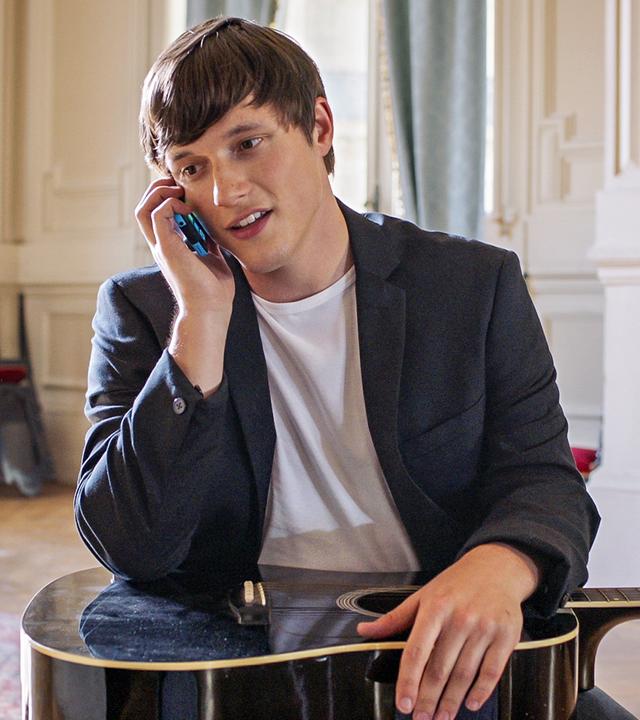 Henri sitzt auf einem Stuhl, seine Gitarre liegt auf seinem Schoß. Mit der rechten Hand hält er sein Handy ans Ohr.
