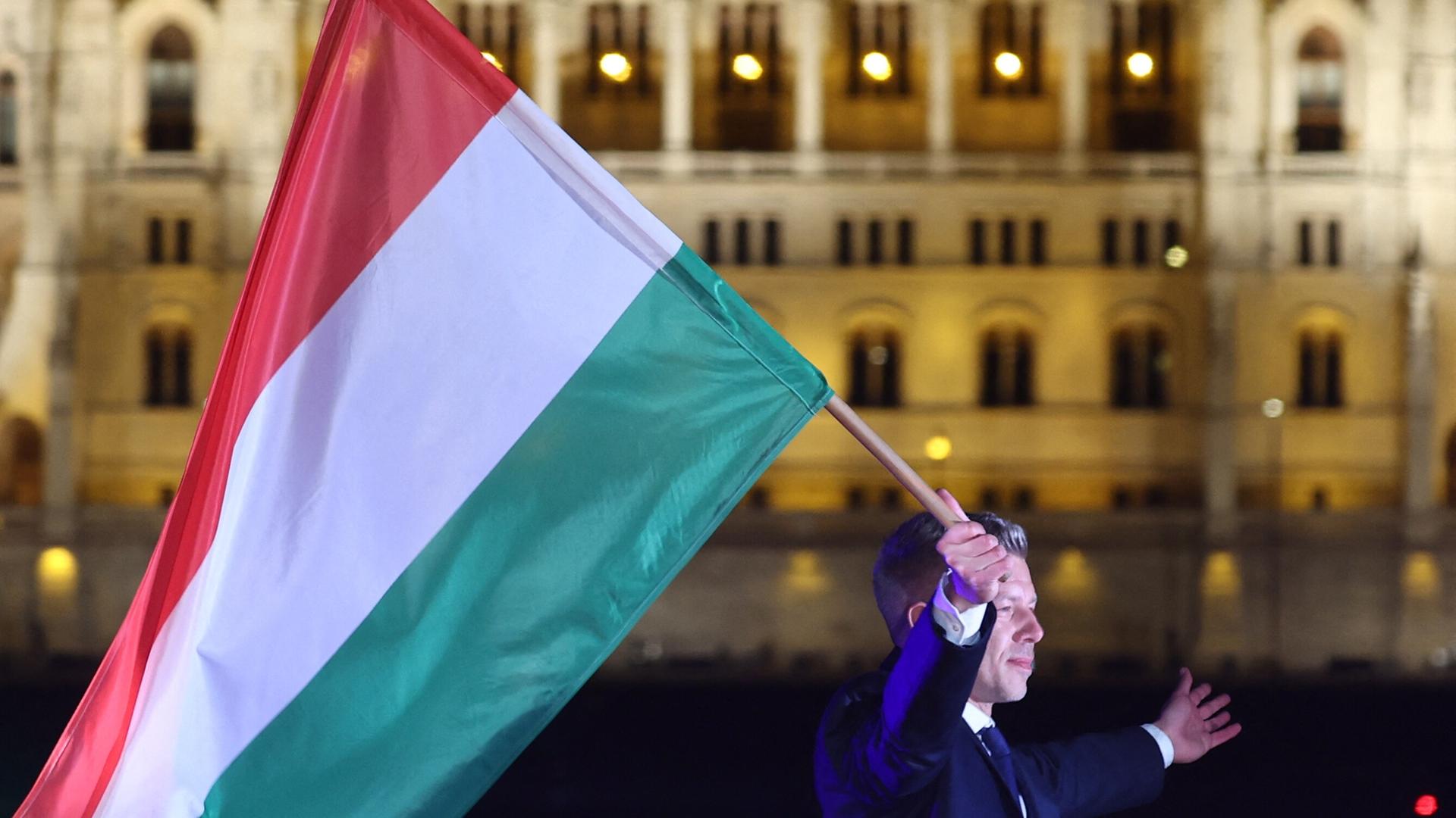 Ein Mann im Anzug läuft mit der ungarischen Flagge im Dunkeln vor einem beleuchteten historischen Gebäude, dem ungarischen Parlament in Budapest.