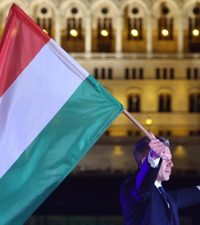 Ein Mann im Anzug läuft mit der ungarischen Flagge im Dunkeln vor einem beleuchteten historischen Gebäude, dem ungarischen Parlament in Budapest.