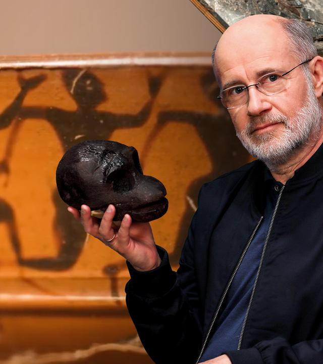 Harald Lesch hält eine schwarze Skulptur eines Schädels in der Hand. Hinter ihm ist eine Wandmalerei zu sehen, die eine Reihe von Frauen in Bewegung zeigt.
