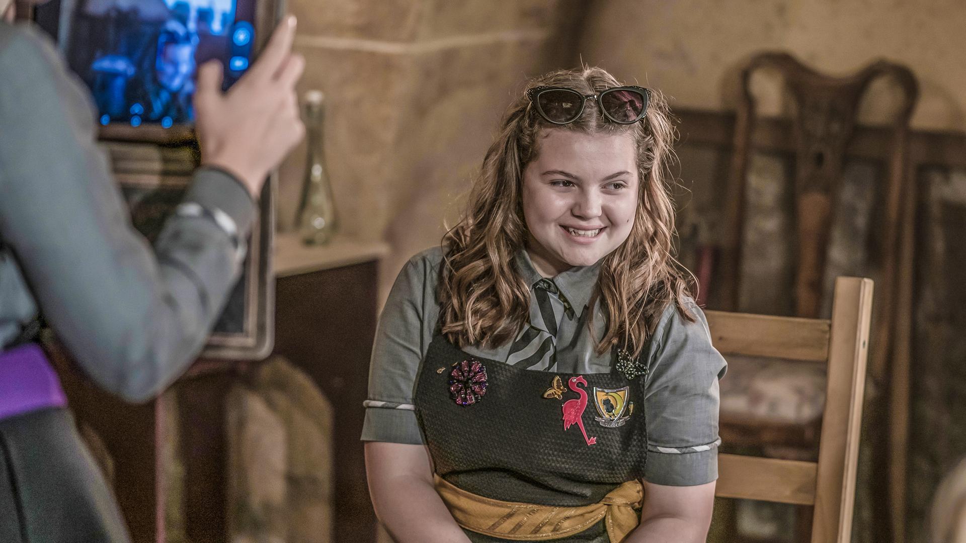 Im Klassenzimmer: Maude sitzt gestylt auf eine Stuhl in Pose. Sie wird von mehreren Mitschülern gefilmt und interviewt.