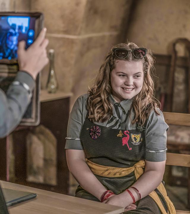 Im Klassenzimmer: Maude sitzt gestylt auf eine Stuhl in Pose. Sie wird von mehreren Mitschülern gefilmt und interviewt.