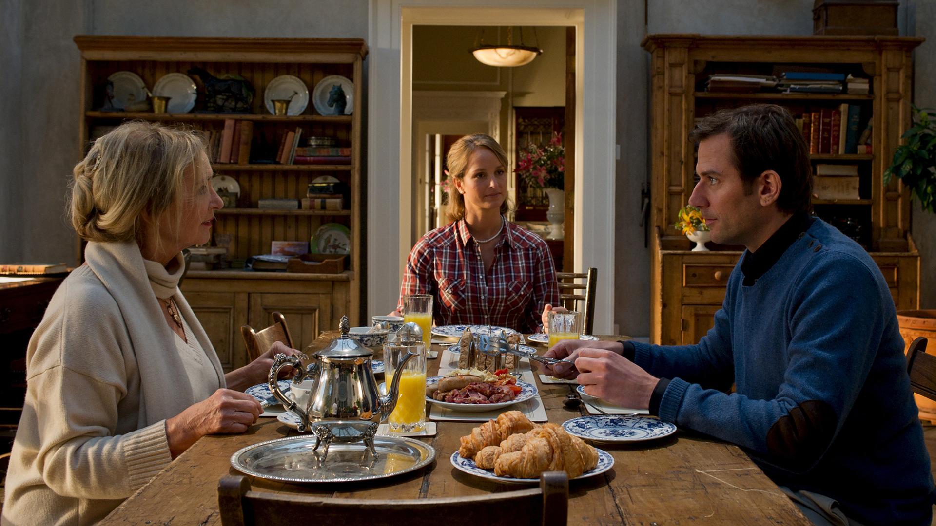 Gillian (Mitte, Hintergrund) sitzt zusammen mit Dylan (rechts, Vordergrund) und dessen Mutter Lady Ava Brooks (links, Vordergrund) am Frühstückstisch.