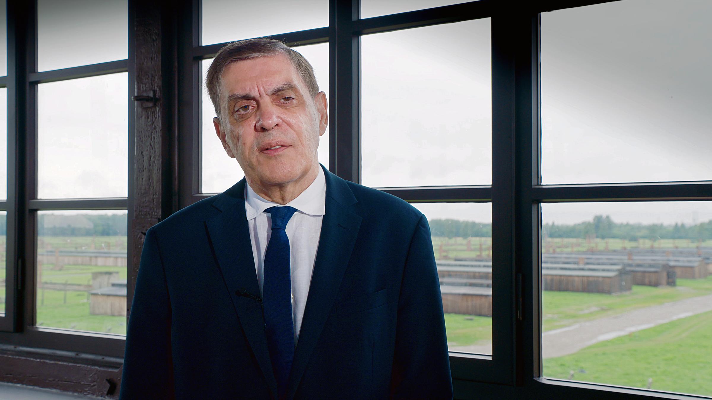 Ein älterer bartloser Mann mit graumelierten kurzen Haaren, der ein dunkelblaues Sakko und einen dunkelblauen Schlips und ein weißes Hemd trägt – Romani Rose -, steht vor einem Fenster im ersten oder zweiten Stock eines Gebäudes und spricht. Hinter ihm hat man die Aussicht über die Überreste des ehemaligen Lagers Auschwitz-Birkenau.