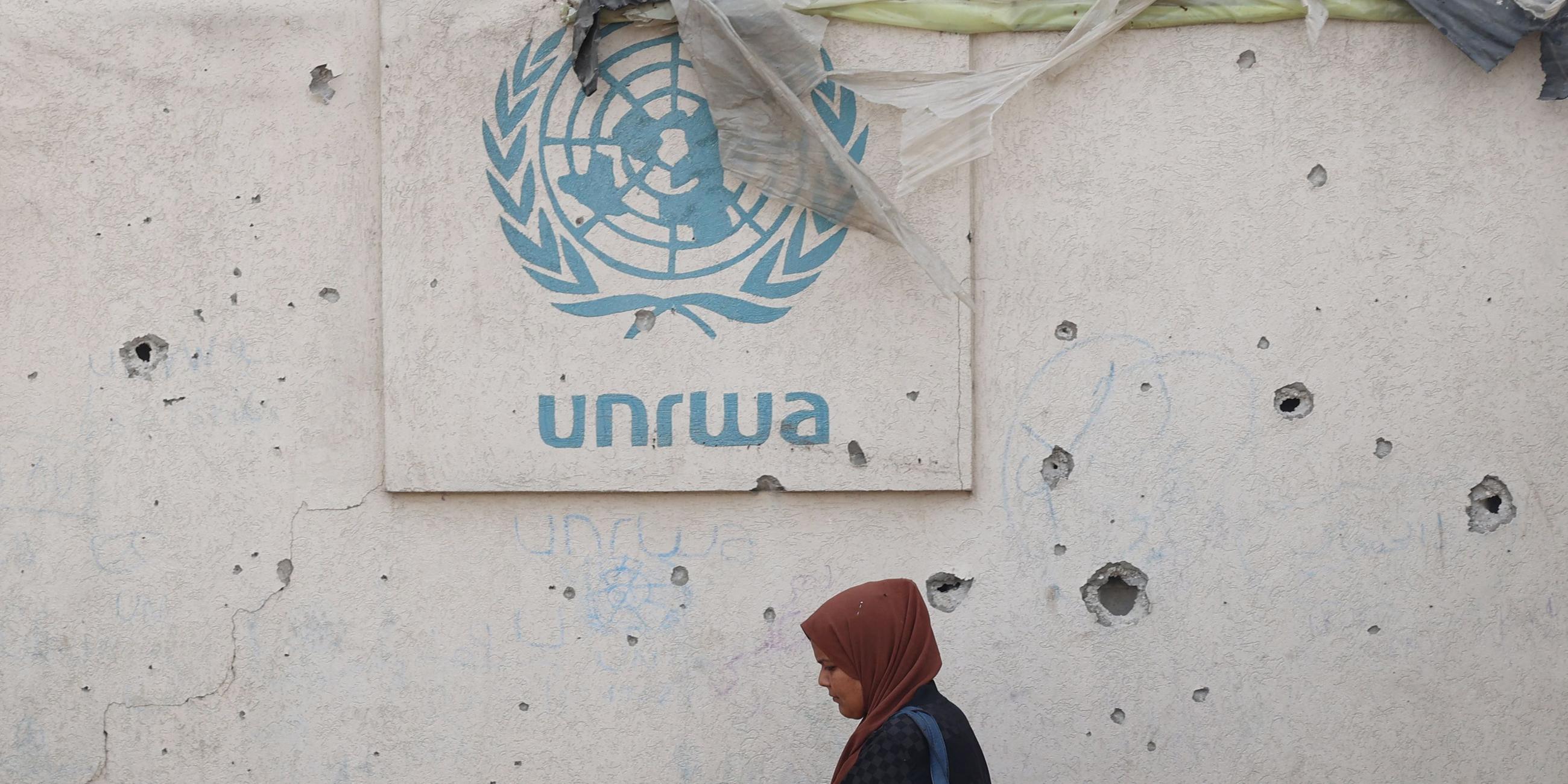 Eine Palästinenserin läuft in einem Lager für Vertriebene in Rafah an einer beschädigten Wand vorbei, daran befestigt ist das Logo des Hilfswerks der Vereinten Nationen für Palästina-Flüchtlinge im Nahen Osten (UNRWA).