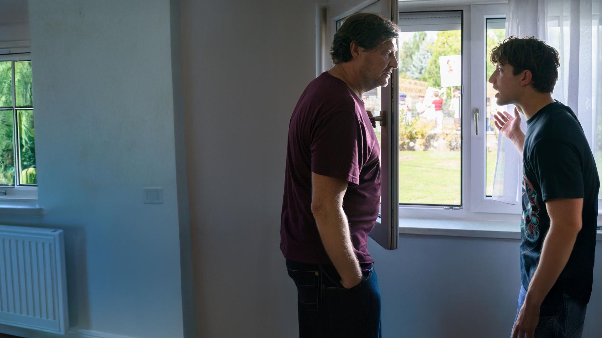 "SOKO Wien - Unschuldsvermutung": Gernot Polmer (Harald Windisch) und Lukas Polmer (Imre Lichtenberger) stehen sich vor einem geöffneten Fenster gegenüber. Sie blicken sich alarmiert an. Lukas Polmer gestikuliert mit seiner Hand nach draußen, wo sich eine Menschenmenge befindet, die mehrere beschriftete und bebilderte Schilder hochhält.