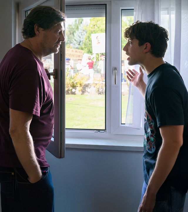 "SOKO Wien - Unschuldsvermutung": Gernot Polmer (Harald Windisch) und Lukas Polmer (Imre Lichtenberger) stehen sich vor einem geöffneten Fenster gegenüber. Sie blicken sich alarmiert an. Lukas Polmer gestikuliert mit seiner Hand nach draußen, wo sich eine Menschenmenge befindet, die mehrere beschriftete und bebilderte Schilder hochhält.