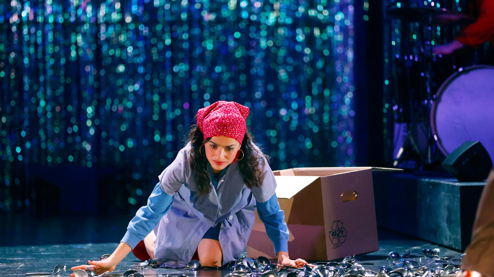 Schauspielerin in hellblauem Arbeitskittel, rotem Kopftuch mit weißen Punkten und blauen Ärmeln kniet auf einer Bühne, umgeben von verstreuten Löffeln. Sie stützt sich mit den Händen ab und blickt besorgt nach unten. Hinter ihr steht ein geöffneter Pappkarton mit Recycling-Symbol. Im Hintergrund ist ein glitzernder Bühnenvorhang zu erkennen, in der rechten oberen Eckw sind Teile eines Schlagzeugs und eines Musikers zu sehen.