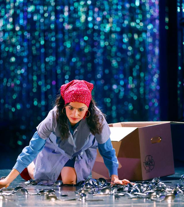 Schauspielerin in hellblauem Arbeitskittel, rotem Kopftuch mit weißen Punkten und blauen Ärmeln kniet auf einer Bühne, umgeben von verstreuten Löffeln. Sie stützt sich mit den Händen ab und blickt besorgt nach unten. Hinter ihr steht ein geöffneter Pappkarton mit Recycling-Symbol. Im Hintergrund ist ein glitzernder Bühnenvorhang zu erkennen, in der rechten oberen Eckw sind Teile eines Schlagzeugs und eines Musikers zu sehen.